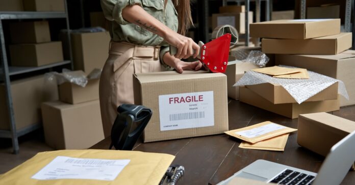 A woman in a green shirt tapes a box labeled fragile amid packing supplies in a room lined with shelves of stored boxes.