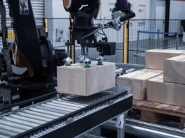 A robotic arm using suction cups to pick up and place a cardboard box on a roller belt in an industry warehouse.