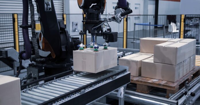 A robotic arm using suction cups to pick up and place a cardboard box on a roller belt in an industry warehouse.
