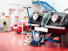 An older car model in a workshop with red flooring with a blue jack raising it into the air on the right side.