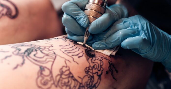 A person who is wearing blue gloves is holding a tattooing needle and is using it one another person.