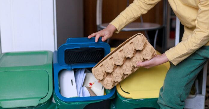 A person leaning in to throw something into the recycling bin. The bins have yellow, blue, and green lids.