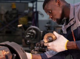 A Black man wearing gloves holds a grooved circular gear while operating a metal lathe machine in a factory.