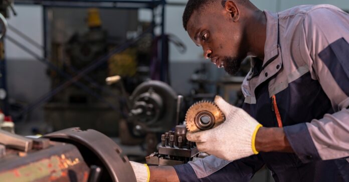 A Black man wearing gloves holds a grooved circular gear while operating a metal lathe machine in a factory.
