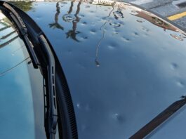 A black car parked on the side of the road with small dents across the hood. These dents are from a hail storm.
