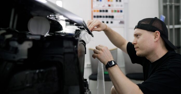 A man wearing a black hat applies a protective vinyl wrap on a black vehicle in an automotive workshop.