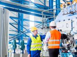 Two workers in safety vests and hard hats inspect large industrial machinery inside a factory with steel beams and tanks.