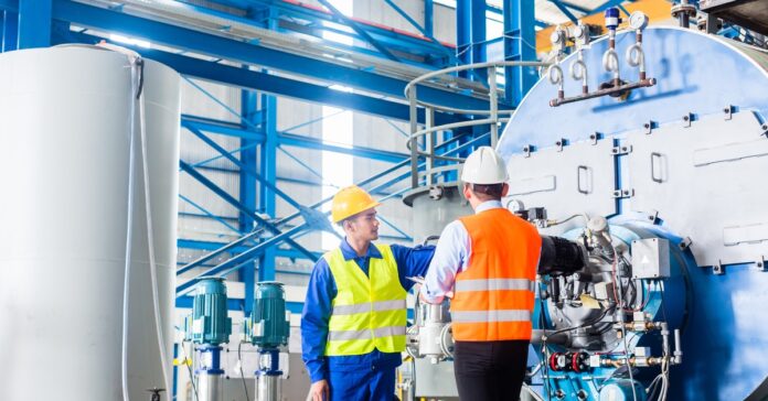 Two workers in safety vests and hard hats inspect large industrial machinery inside a factory with steel beams and tanks.
