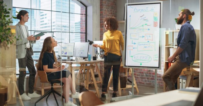 A woman wearing a yellow shirt presents a project timeline using a whiteboard to her fellow colleagues.