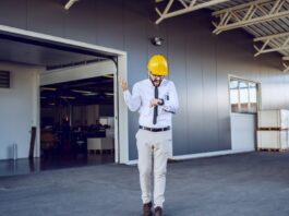 A man in business attire and a hard hat looking down at his watch and yelling. He is walking away from a building.