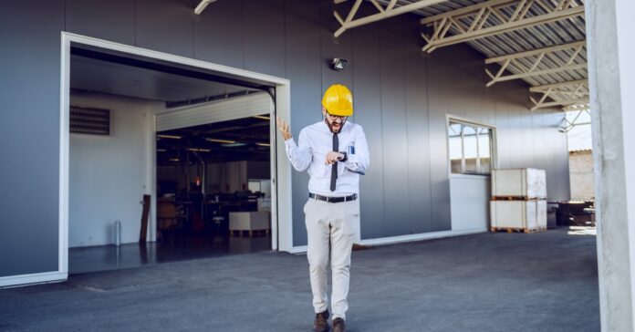A man in business attire and a hard hat looking down at his watch and yelling. He is walking away from a building.