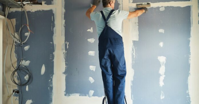 A man wearing blue overalls is standing on a step ladder while taping drywall. The wall is blue with white patches.