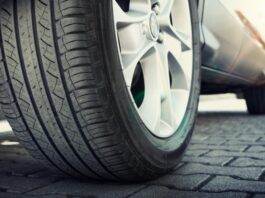 A ground-level view of the tire of a car with deep tread and a silver rim. The vehicle is parked on a road with gray pavers.