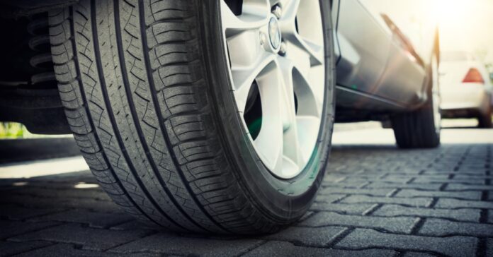 A ground-level view of the tire of a car with deep tread and a silver rim. The vehicle is parked on a road with gray pavers.