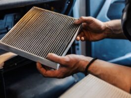 A mechanic holds a dirty cabin filter from a vehicle. A new, clean filter is lying in the foreground.