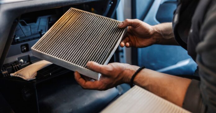 A mechanic holds a dirty cabin filter from a vehicle. A new, clean filter is lying in the foreground.