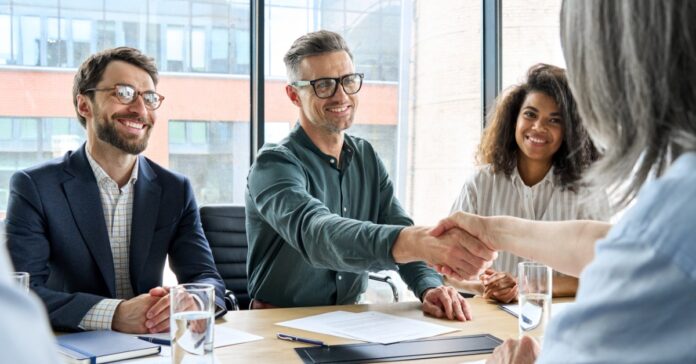 Three smiling businesspeople sit on one side of a table. One of them is shaking hands with someone across the table.