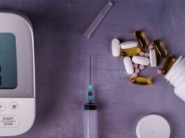 A blood pressure monitor, an uncapped needle, and a bottle tipped over with different medications spilling out on a gray table.