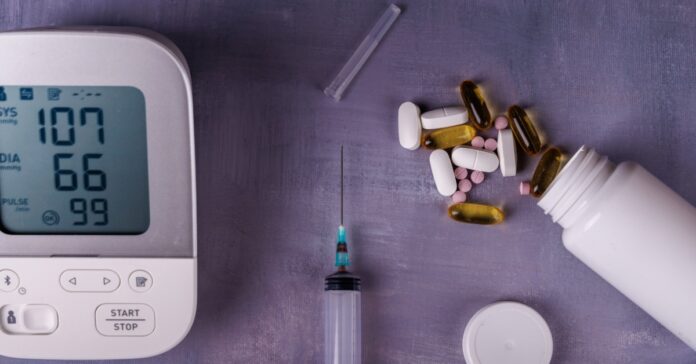 A blood pressure monitor, an uncapped needle, and a bottle tipped over with different medications spilling out on a gray table.