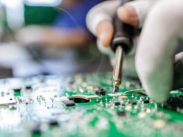 A gloved technician uses a soldering iron on a green circuit board in a close-up shot of a manufacturing process.