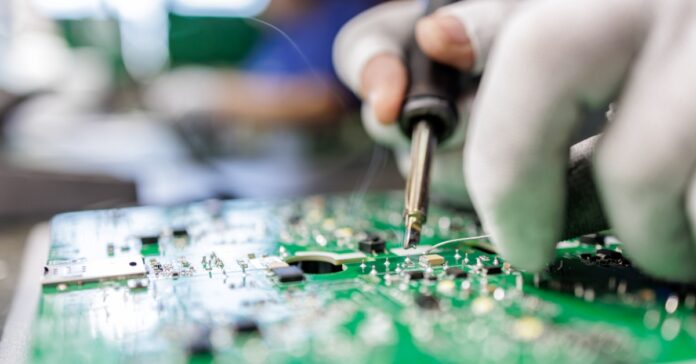 A gloved technician uses a soldering iron on a green circuit board in a close-up shot of a manufacturing process.