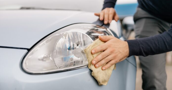 A close-up of a person in a dark long-sleeve shirt using a cloth to clean the headlight of a light-colored car.