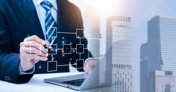A man in a suit touching a box in a workflow process graphic in front of his laptop with a gradient of skyscrapers.