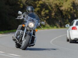 A motorcycle rider wearing a helmet, leather jacket, and gloves while riding their motorcycle on a road near a car.
