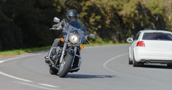 A motorcycle rider wearing a helmet, leather jacket, and gloves while riding their motorcycle on a road near a car.