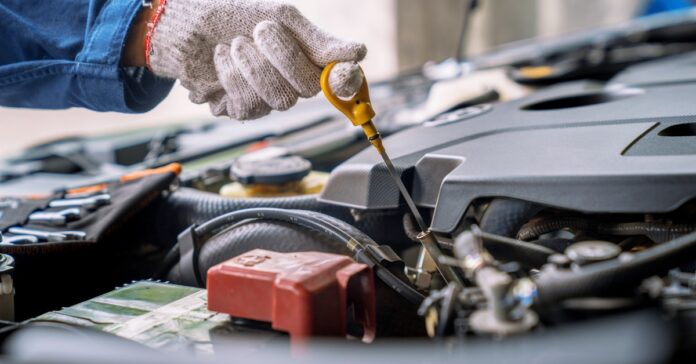 A person working under an open car hood in a garage service bay while wearing coveralls and a pair of gloves.