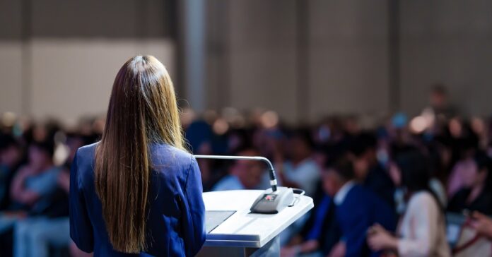 A woman standing at a podium in front of a large audience. There is a mic on the stand and a notebook.