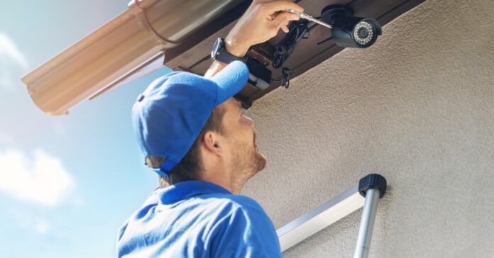 A man in a blue shirt and cap stands on a ladder and uses a screwdriver to install a security camera on the side of a home.