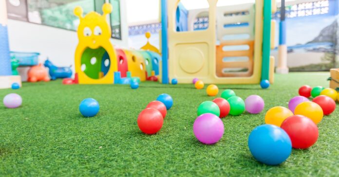 Colorful plastic balls scattered across artificial grass in a bright indoor playground with soft play structures and tunnels in the background.