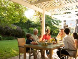 Kids and adults sit for a meal at a patio table under a pergola. The lawn is green, and shrubs line the fence.