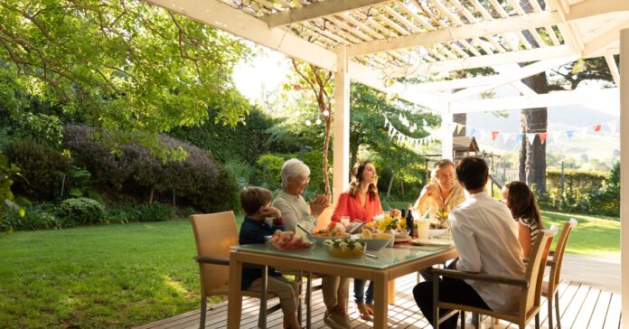 Kids and adults sit for a meal at a patio table under a pergola. The lawn is green, and shrubs line the fence.