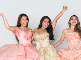 Three girls in a pink, peach, and champagne ball gown stand next to one another and smile with an arm up.