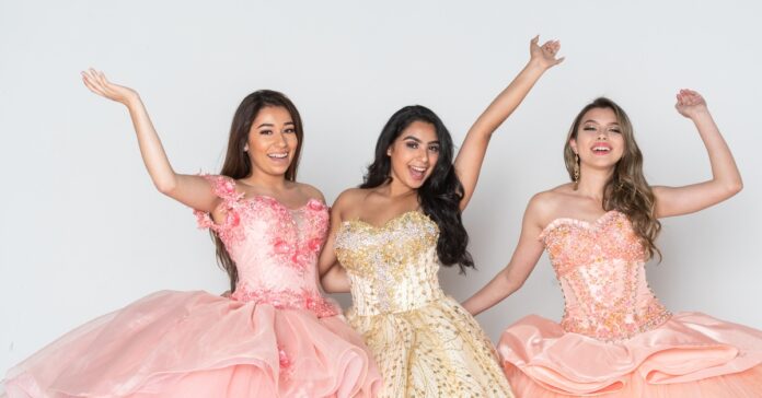 Three girls in a pink, peach, and champagne ball gown stand next to one another and smile with an arm up.