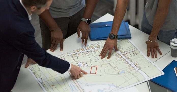 Three men gather around a table, engaged in discussion as they examine a large blueprint of a building plan.