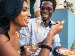 Two people sitting outside in Italy while eating a plate of pasta. The woman is holding a fork and a glass of wine.