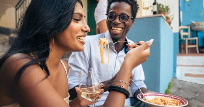 Two people sitting outside in Italy while eating a plate of pasta. The woman is holding a fork and a glass of wine.