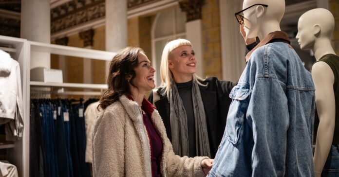 Two women smiling while examining a mannequin dressed in a denim jacket as part of a retail clothing store display.