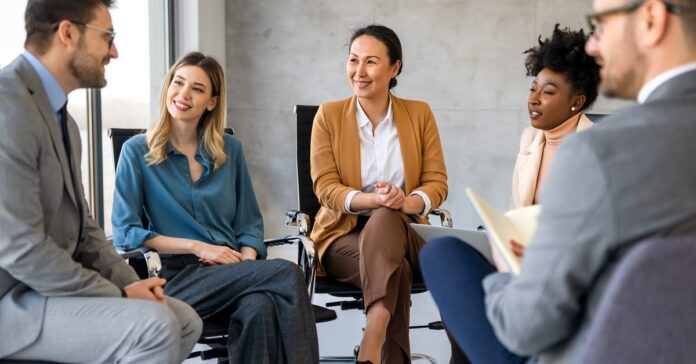 A group of diverse businesspeople sitting in chairs together. They are all looking at one of the men in the circle.