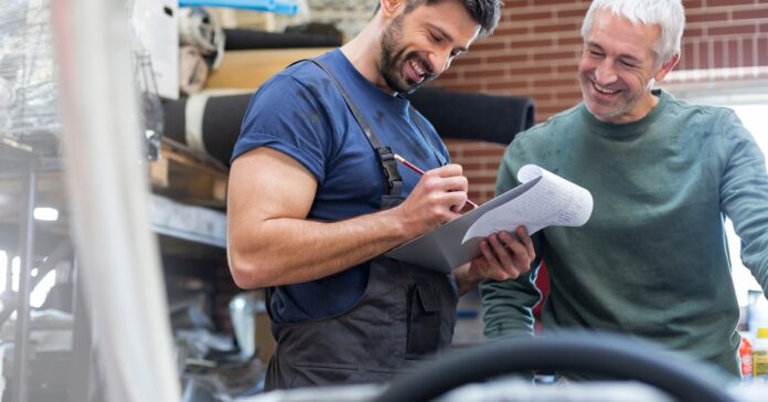 A mechanic in an auto shop smiles as he writes on a clipboard and serves an equally happy-looking customer.