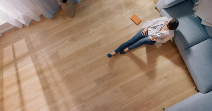A person sitting next to a small book on a wooden floor at home. The person sits against a long gray couch.
