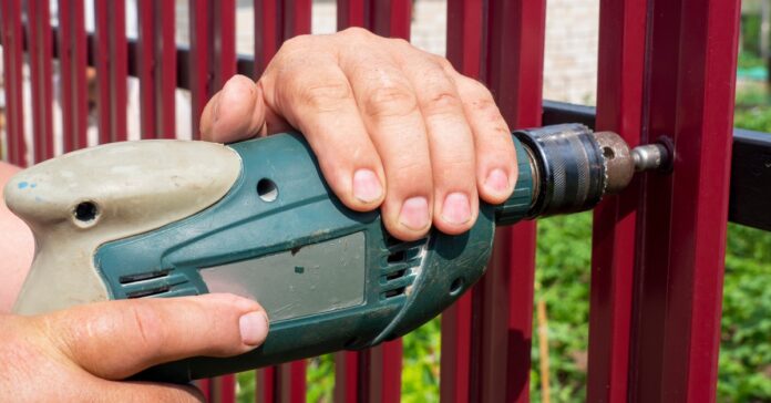 A person using a power drill to fasten a screw into a dark red metal fence panel with green grass behind it.
