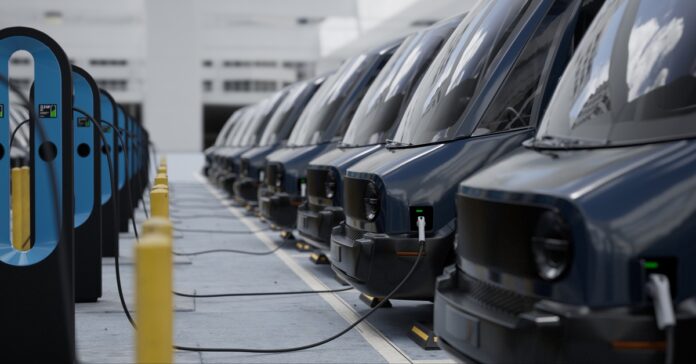 A row of black electric delivery vans charges at docking stations in a commercial fleet parking facility.