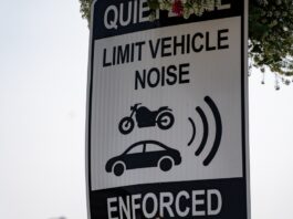 A white sign with black text reading “QUIET ZONE: LIMIT VEHICLE NOISE ENFORCED” and simple black images of a motorcycle and sedan.