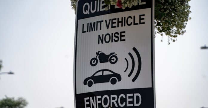 A white sign with black text reading “QUIET ZONE: LIMIT VEHICLE NOISE ENFORCED” and simple black images of a motorcycle and sedan.
