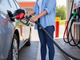 A woman in jeans and a blue overshirt holds a gas nozzle to the fuel tank of her modern silver car at a gas pump.