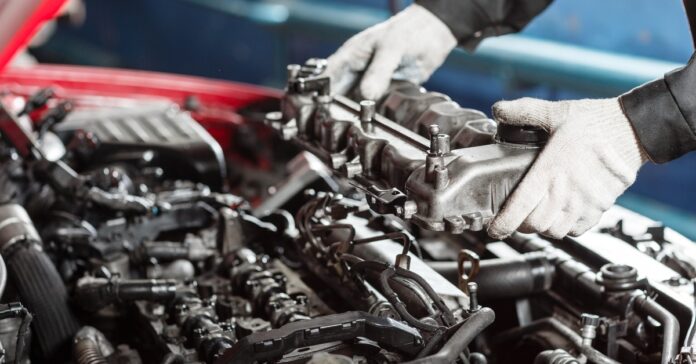 Gloved hands lift a metal engine part above the engine bay under the raised hood of a red vehicle.